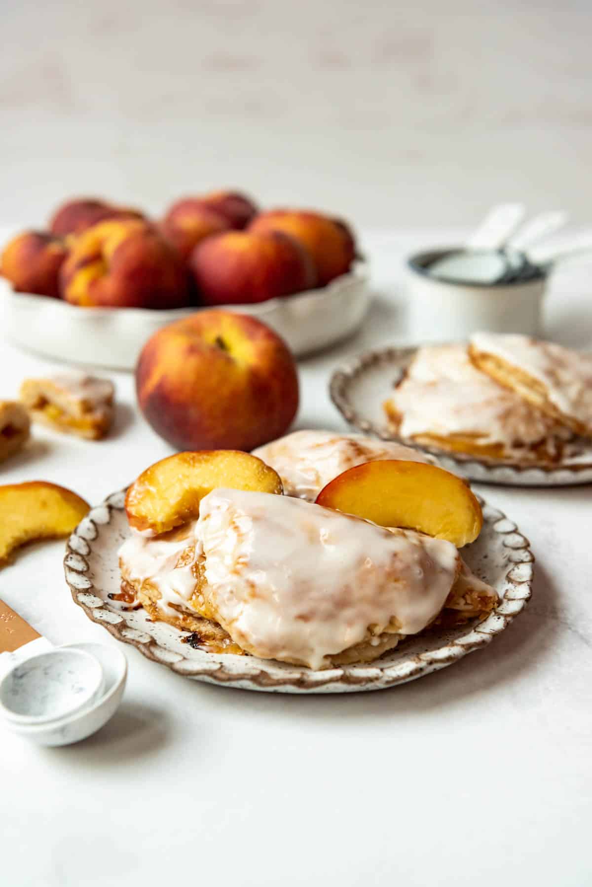 Glazed peach hand pies on a plate with a bowl of peaches behind them.