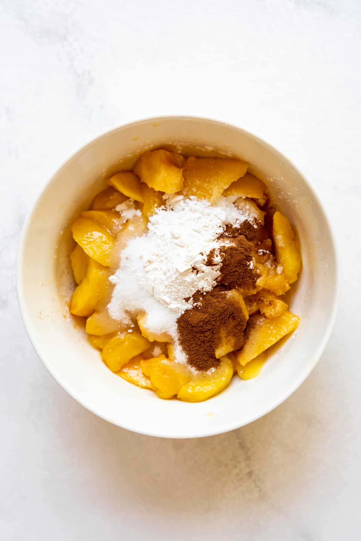 Combining sliced fresh peaches with sugar, cinnamon, and cornstarch in a white bowl.
