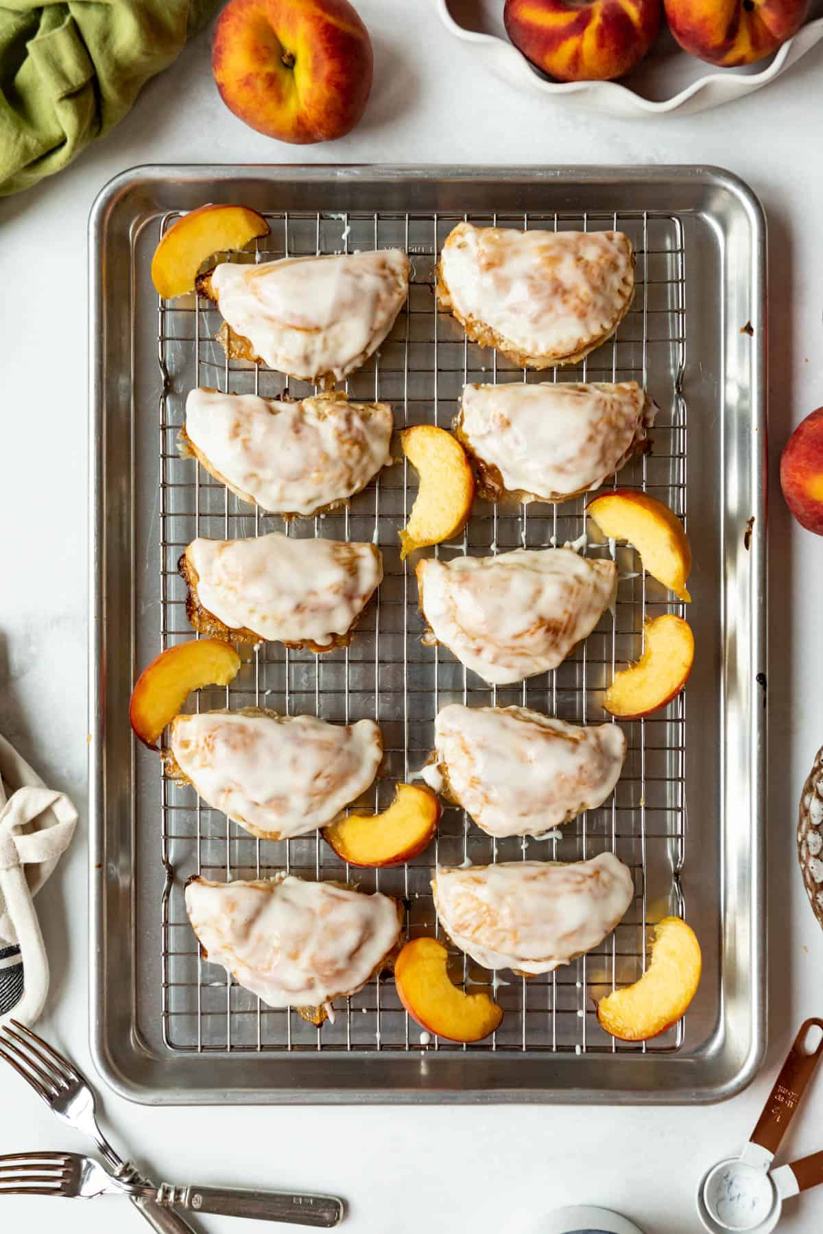 Baked and glazed peach hand pies on a baking sheet lines with a wire rack next to slices of fresh peaches.