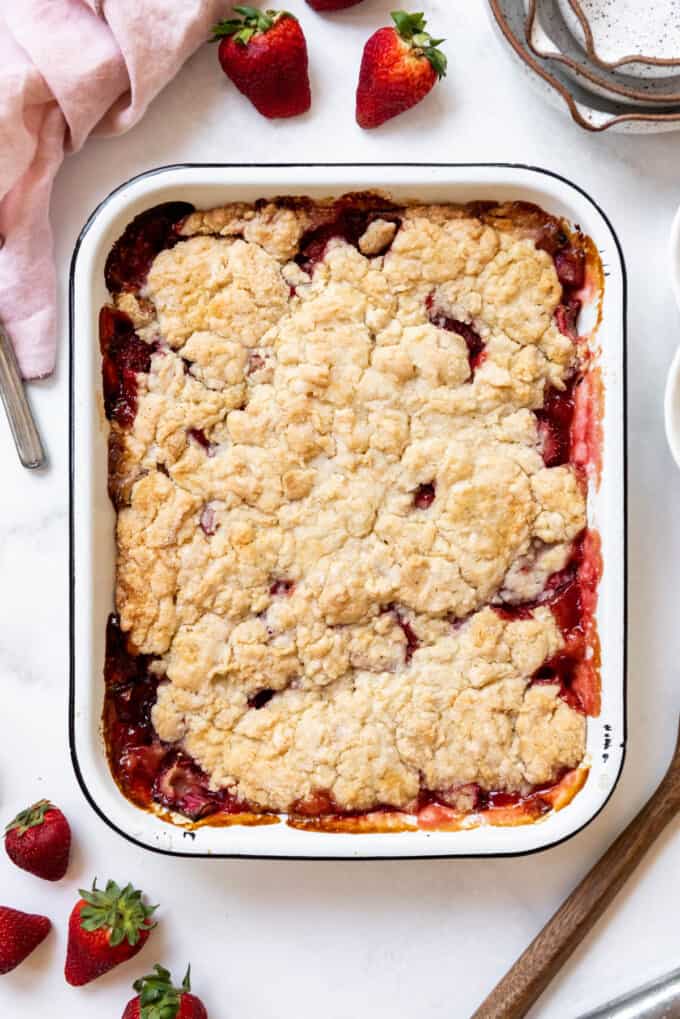 Baked strawberry rhubarb cobbler surrounded by strawberries and a pink cloth napkin.