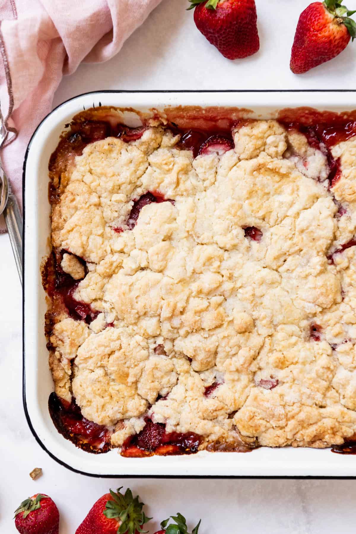 A homemade strawberry rhubarb cobbler with a few strawberries scattered nearby.