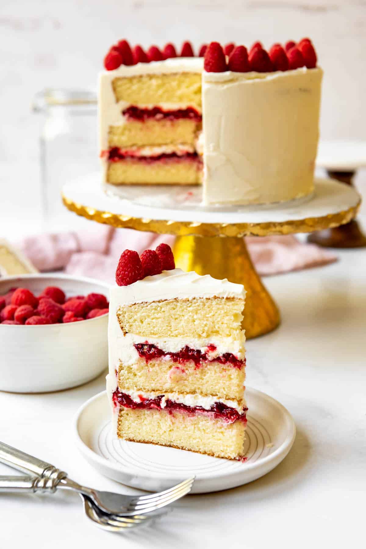 A slice of white chocolate raspberry cake standing on a plate in front of the rest of the cake on a cake stand.