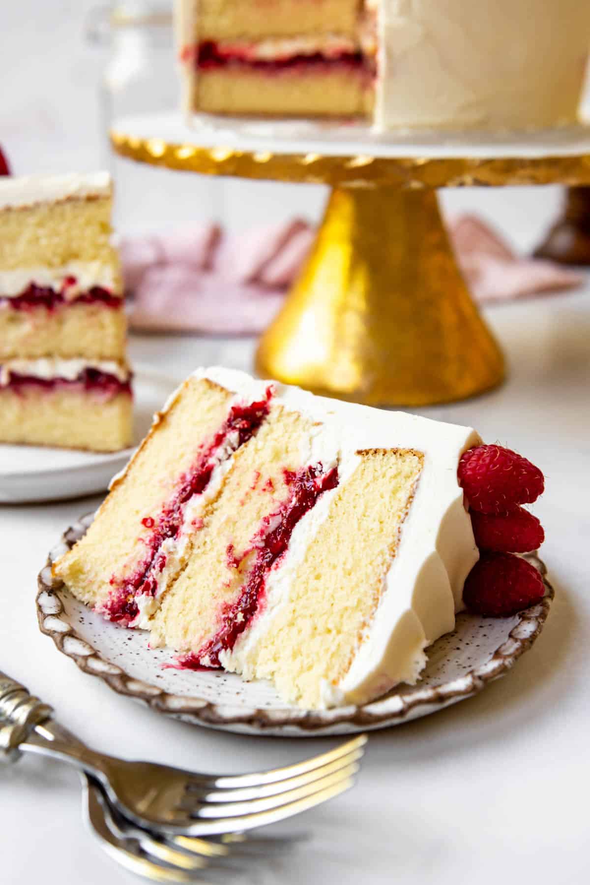 A slice of white chocolate raspberry cake resting on its side on a plate. Other slices and the remaining cake are visible in the background. 