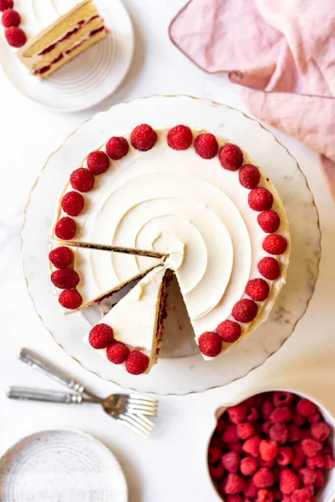 An overhead image of a cake decorated with white frosting and whole raspberries with some slices removed.