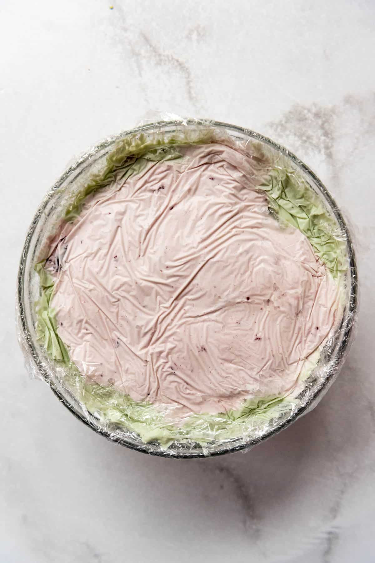 A glass bowl filled with cherry and pistachio ice cream, covered in plastic wrap.