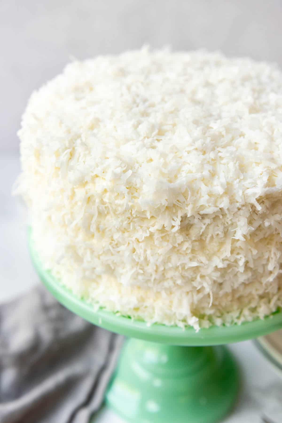 Close up of a green cake stand with a frosted and coconut-covered cake on top.