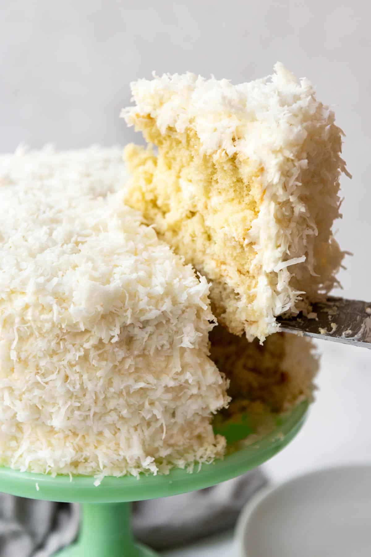 Close up of a green cake stand with a frosted and coconut-covered cake on top with a slice of cake being lifted out of it with a cake slice.