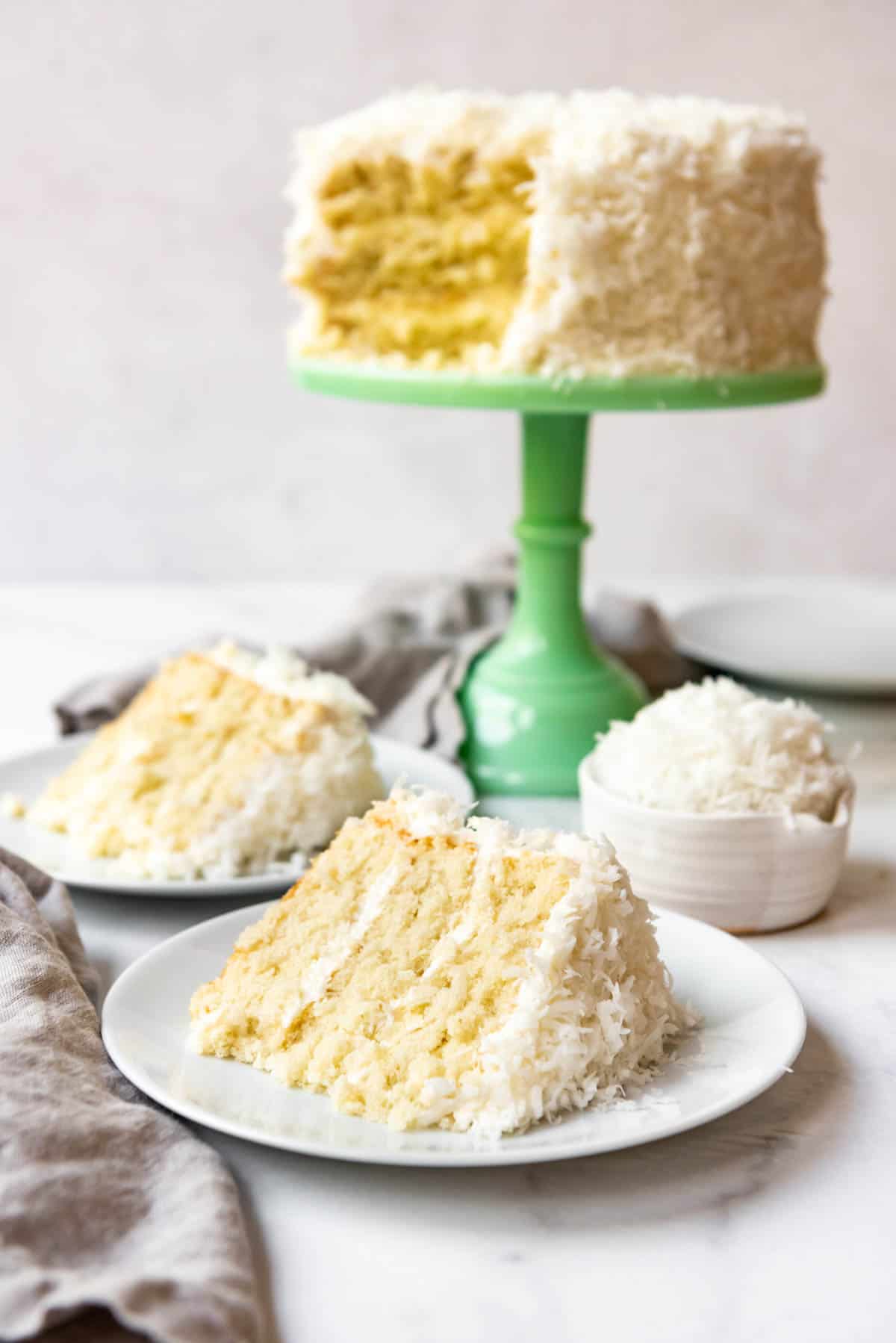 Two plates with slices of Coconut cake on them next to a green cake stand with the rest of the coconut cake on it.