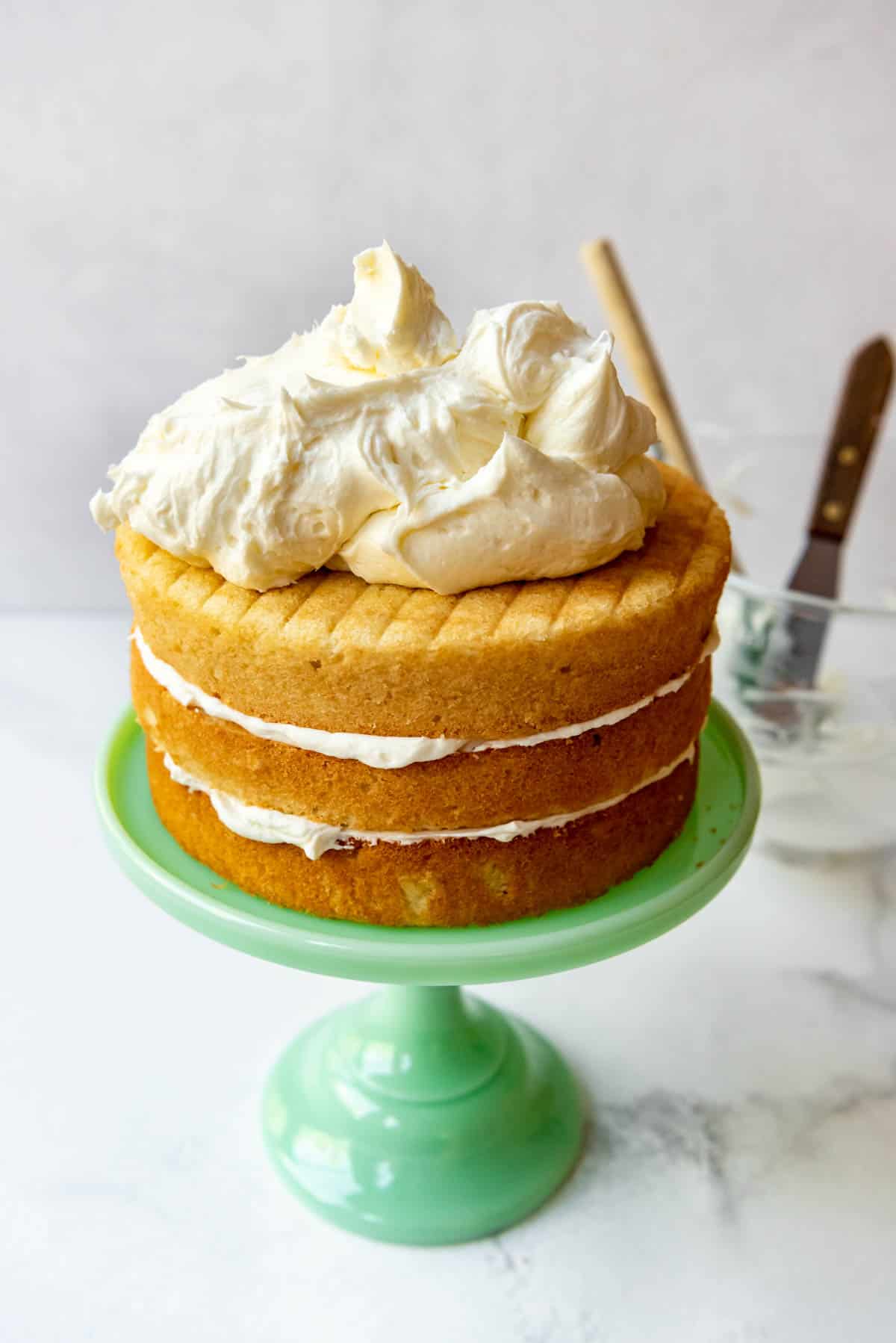A green cake stand with a three-tiered coconut cake on top, with frosting between each layer and a heaped pile of frosting on top, ready to be smoothed out.