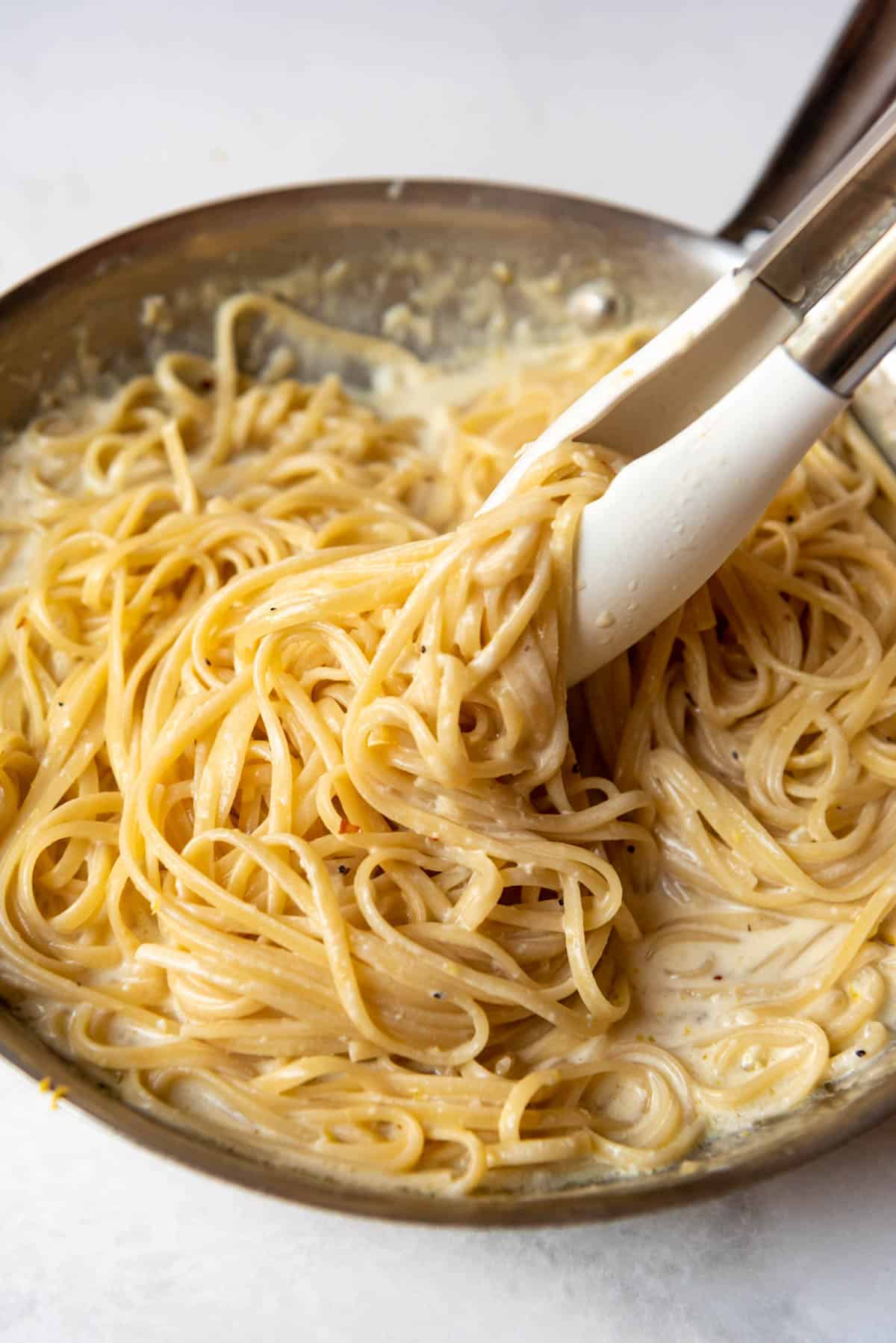 Tossing pasta with tongs in a creamy lemon garlic alfredo sauce.