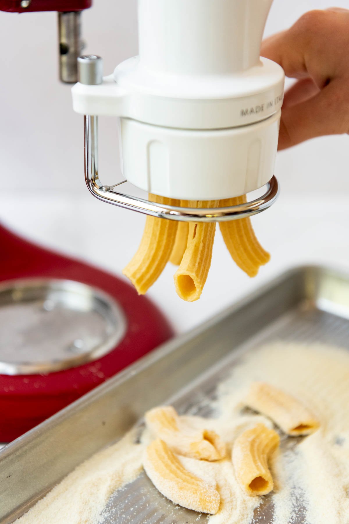Fresh rigatoni pasta tubes being extruded from a KitchenAid pasta attachment.