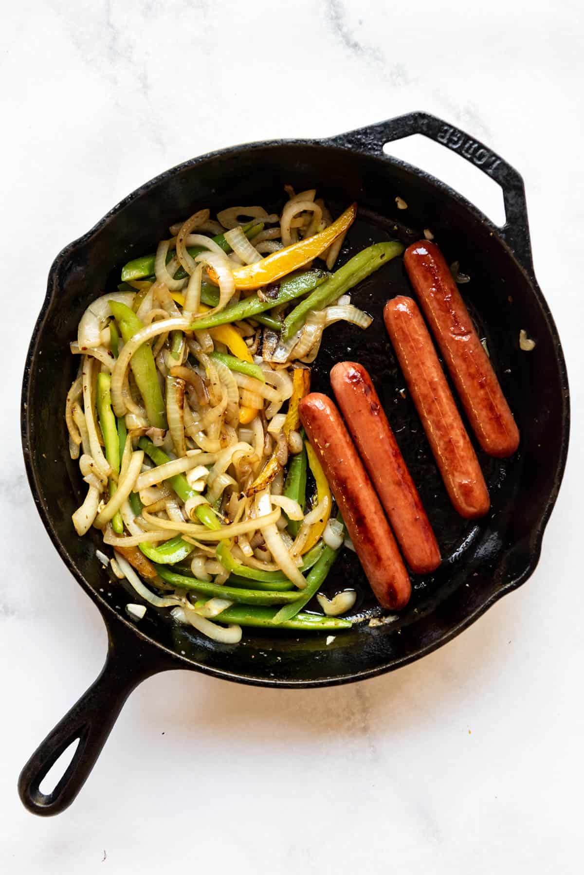 A large cast iron skillet with hot dogs and vegetables in it.