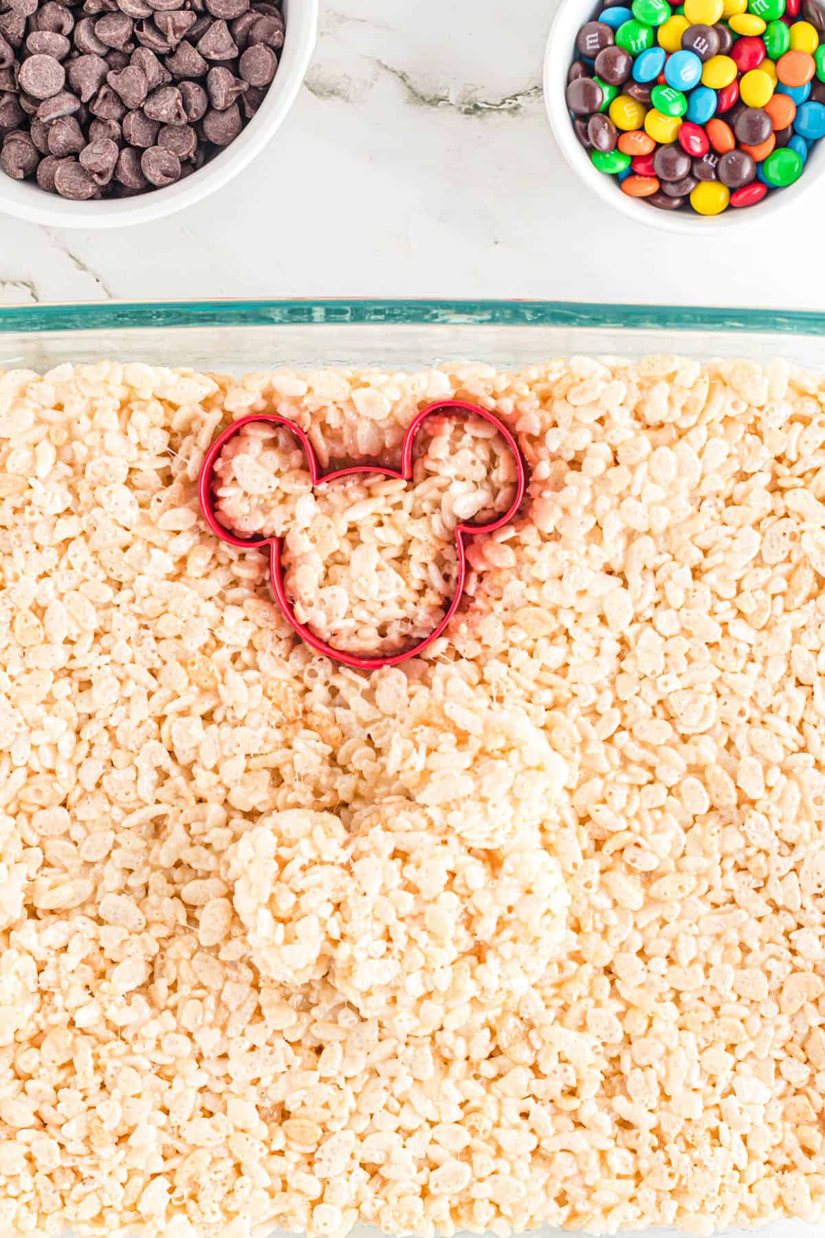 A red Mickey shaped cookie cutter being used to cut out Mickey rice krispies treats.