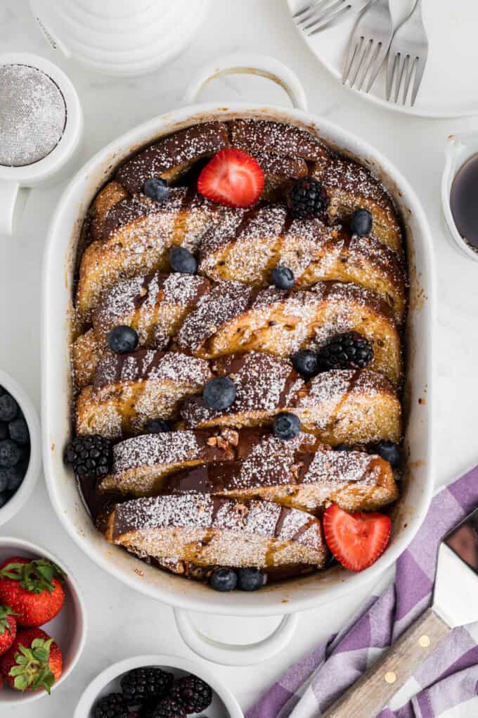 A baking dish over baked french toast drizzled with syrup with fresh fruit.