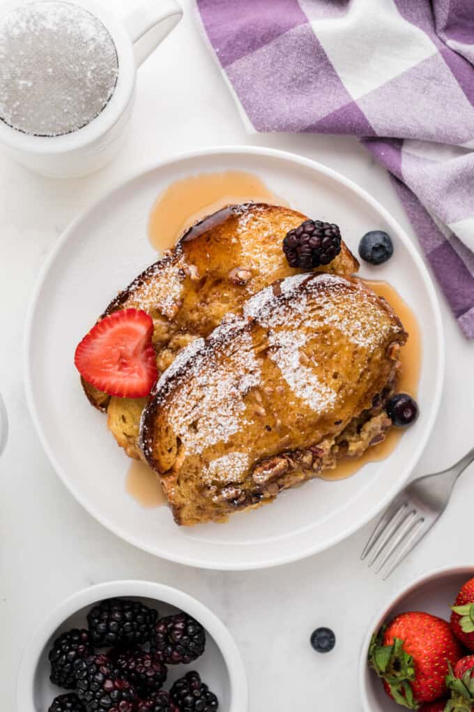 Slices of baked overnight French toast on a white plate with syrup and fresh berries.