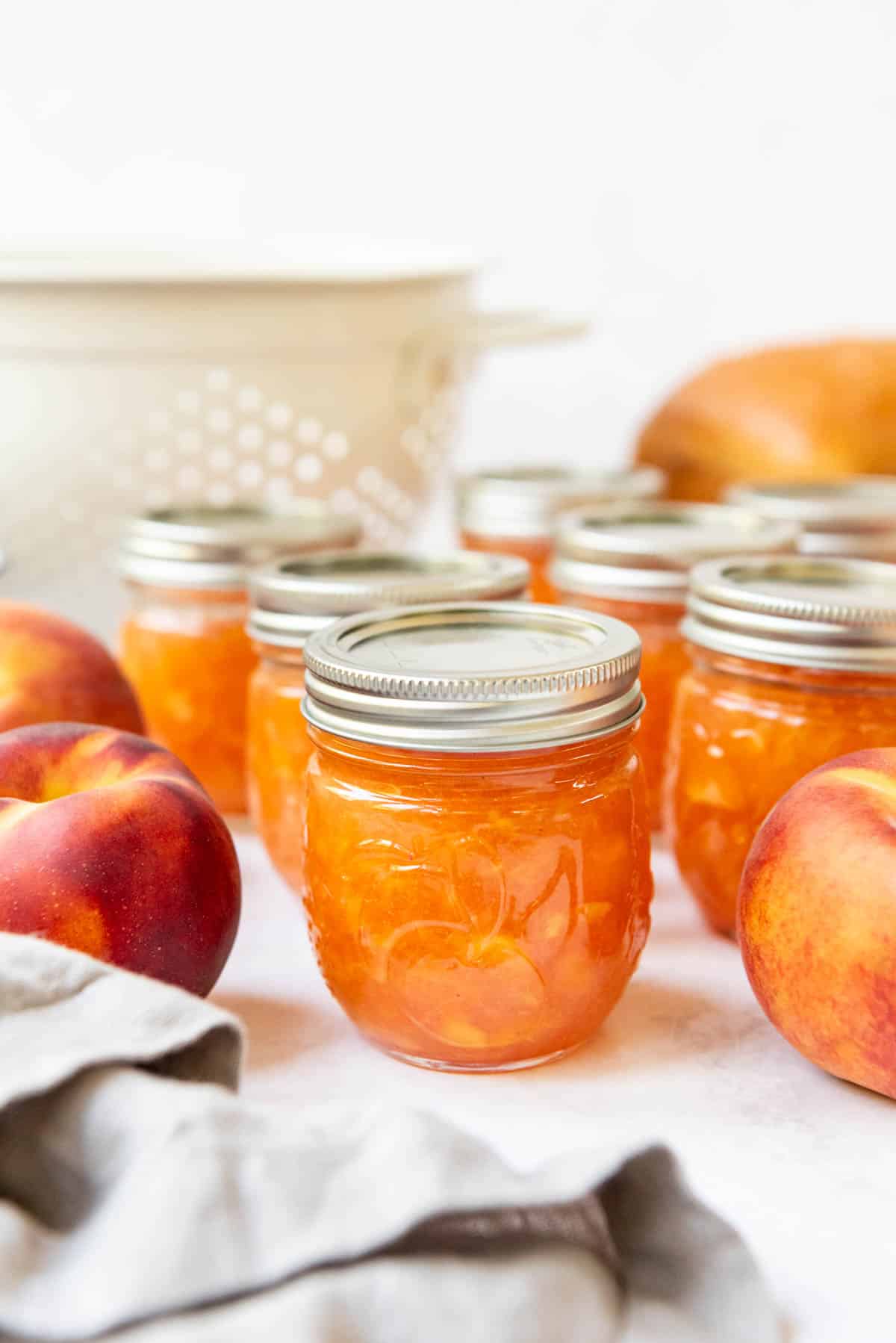 Jars of homemade peach jam in front of a white colander and fresh peaches.