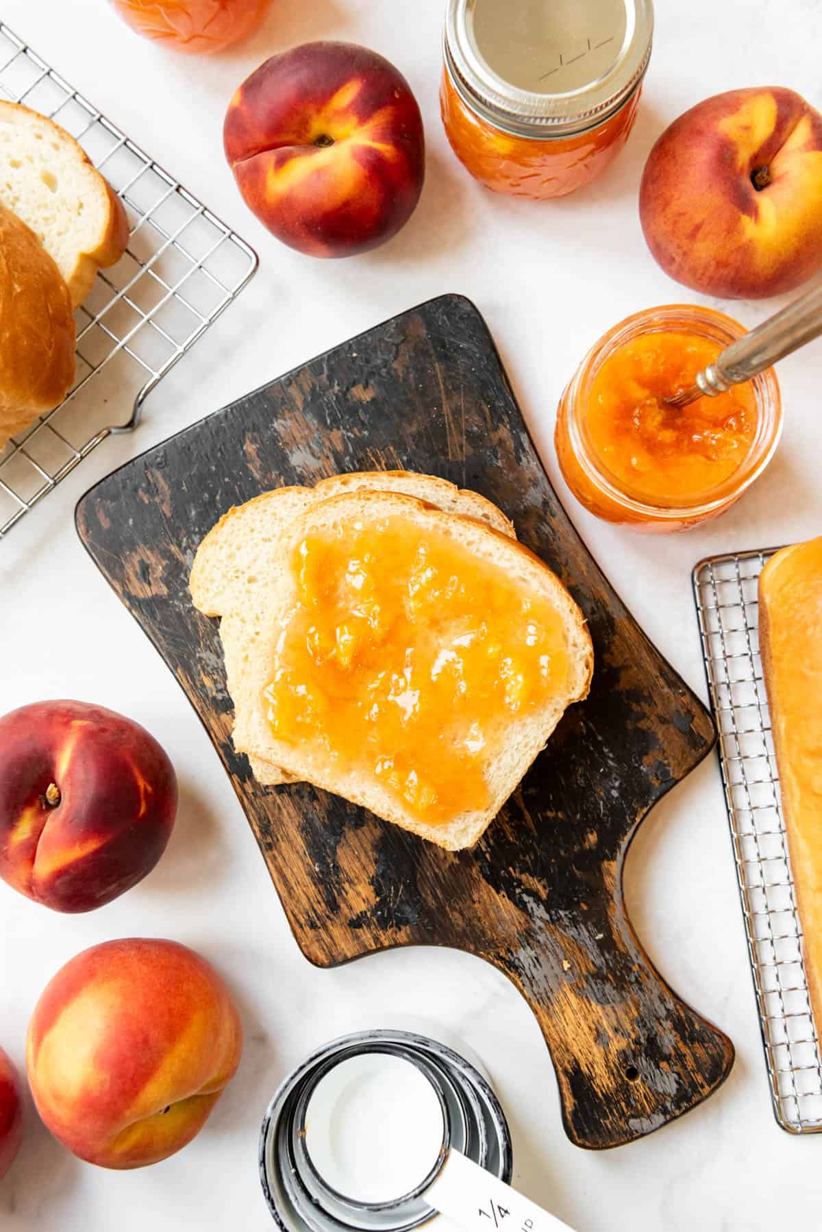 An overhead image of slices of bread with peach jam on a cutting board surrounded by fresh peaches and jars of jam.