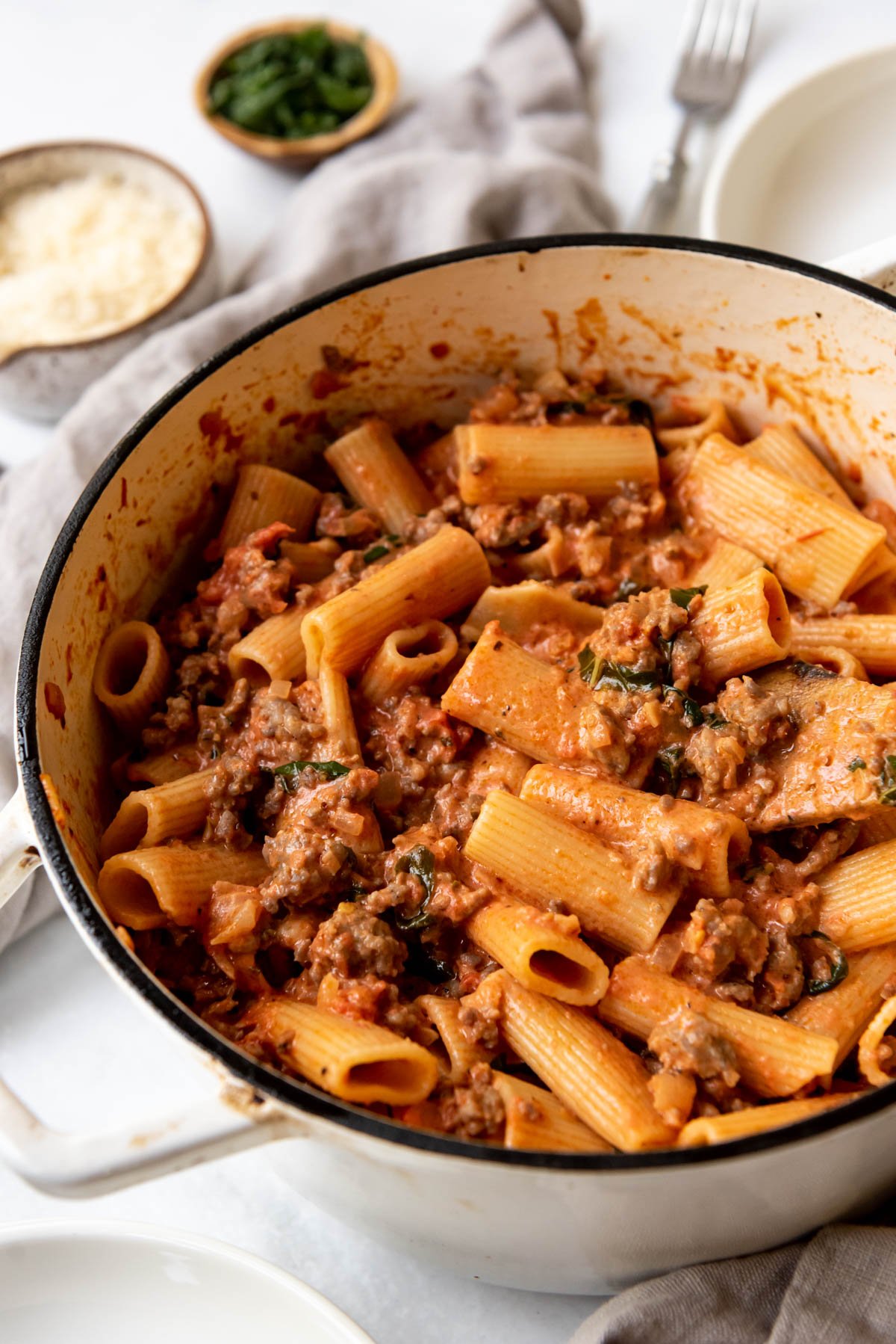 A pot of sausage rigatoni with small bowls of parmesan cheese and fresh basil behind it.