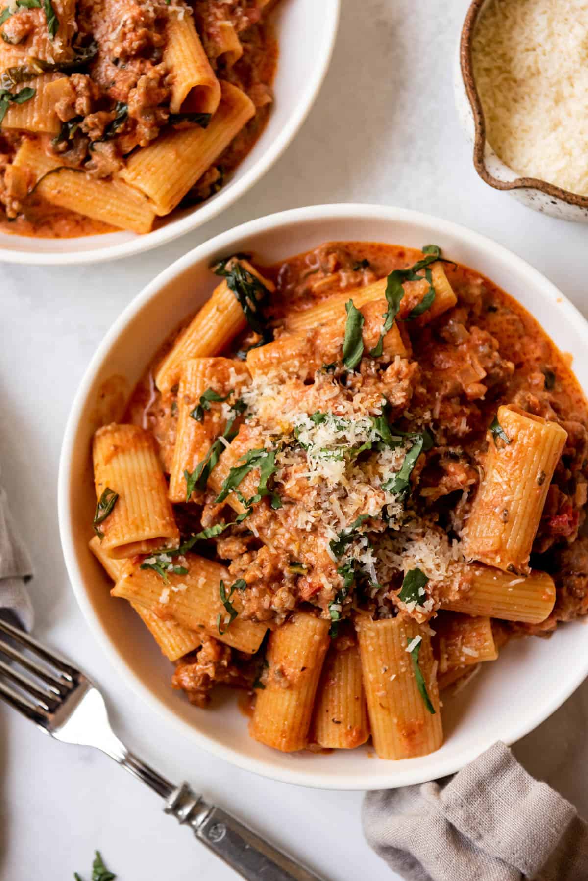 A close image of a bowl of sausage rigatoni with thinly sliced basil and freshly grated parmesan cheese sprinkled on top.