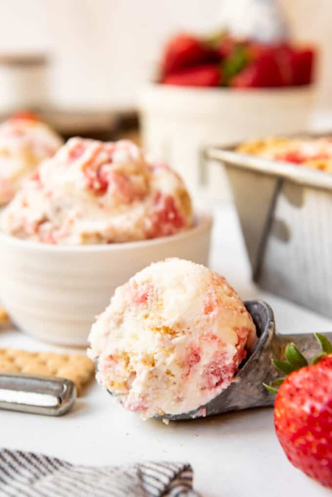 A white bowl filled with Strawberry Cheesecake Ice Cream next to an ice cream scoop with a scoop of the ice cream in it.