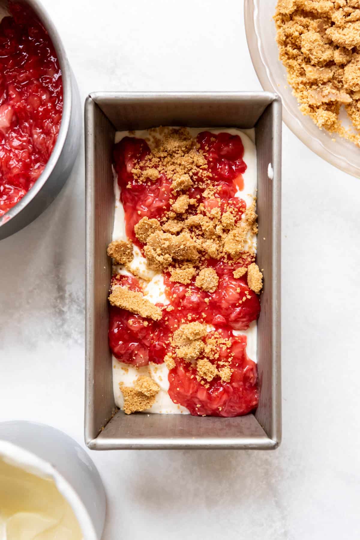Top view of a metal pan half filled with ice cream, strawberry sauce, and graham cracker crumbs.