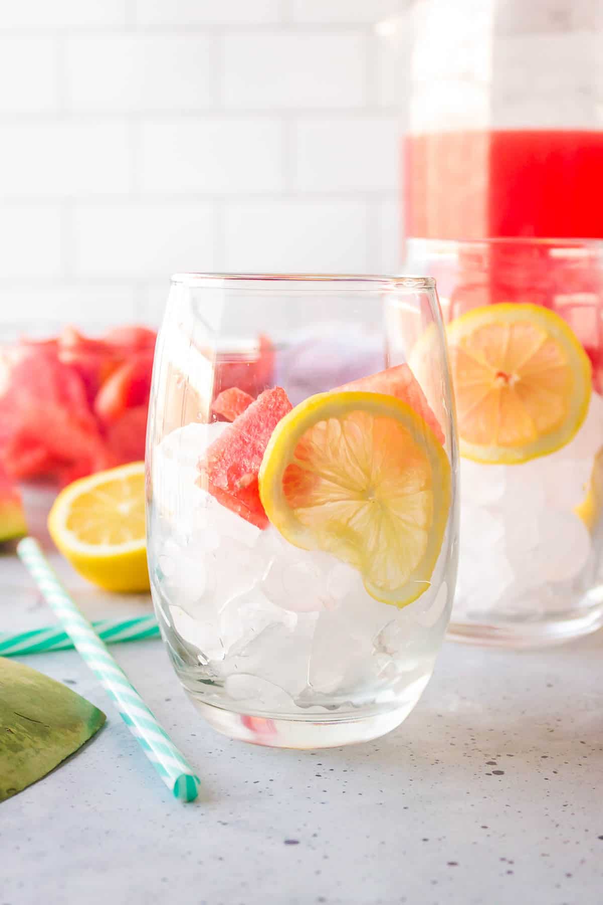A glass with ice, thinly sliced lemonade, and a watermelon chunk.