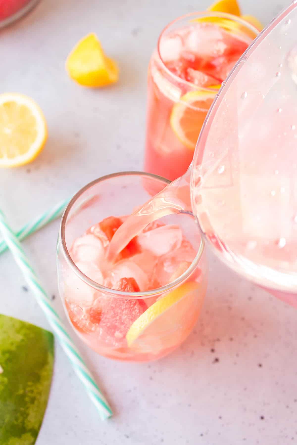 Pouring watermelon lemonade into a glass of ice, watermelon cubes, and lemon slices.
