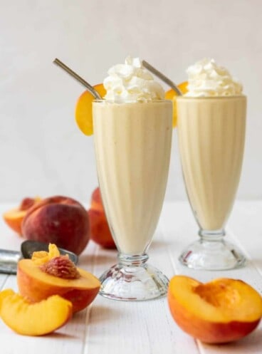 Two peach milkshakes in tall glasses surrounded by fresh peaches.