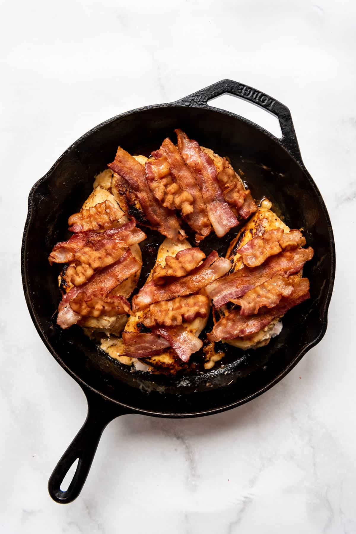 Adding slices of crispy cooked bacon on top of chicken breasts in a cast iron skillet.