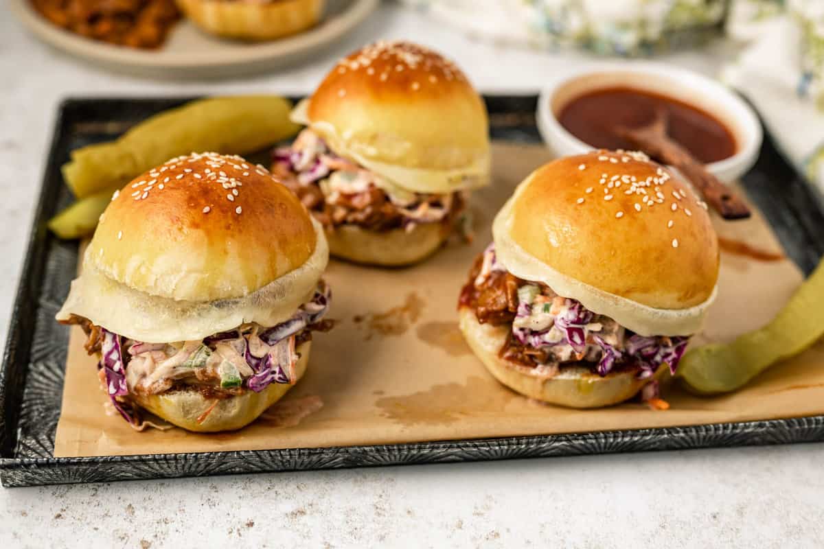 Three bbq beef brisket sandwiches on a baking sheet next to a bowl of barbecue sauce and pickles.