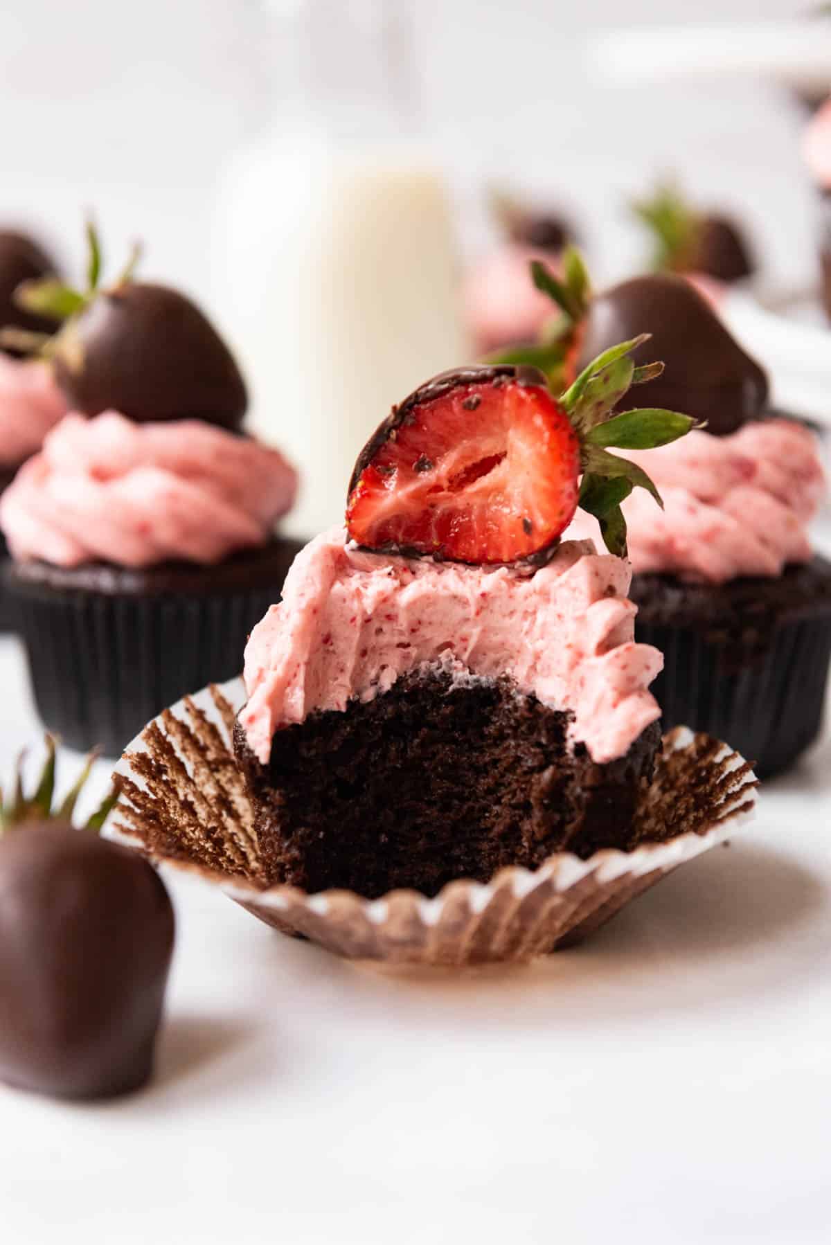 A moist chocolate cupcake with strawberry buttercream frosting and a chocolate covered strawberry on top with a bite taken out of the whole thing.