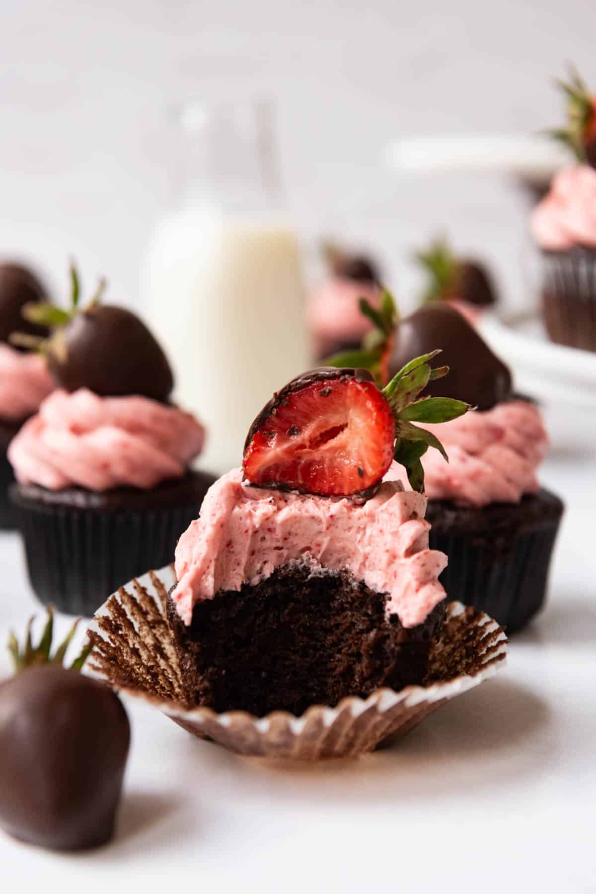 A chocolate covered strawberry cupcake with a bite taken out of it in front of more cupcakes and a bottle of milk.