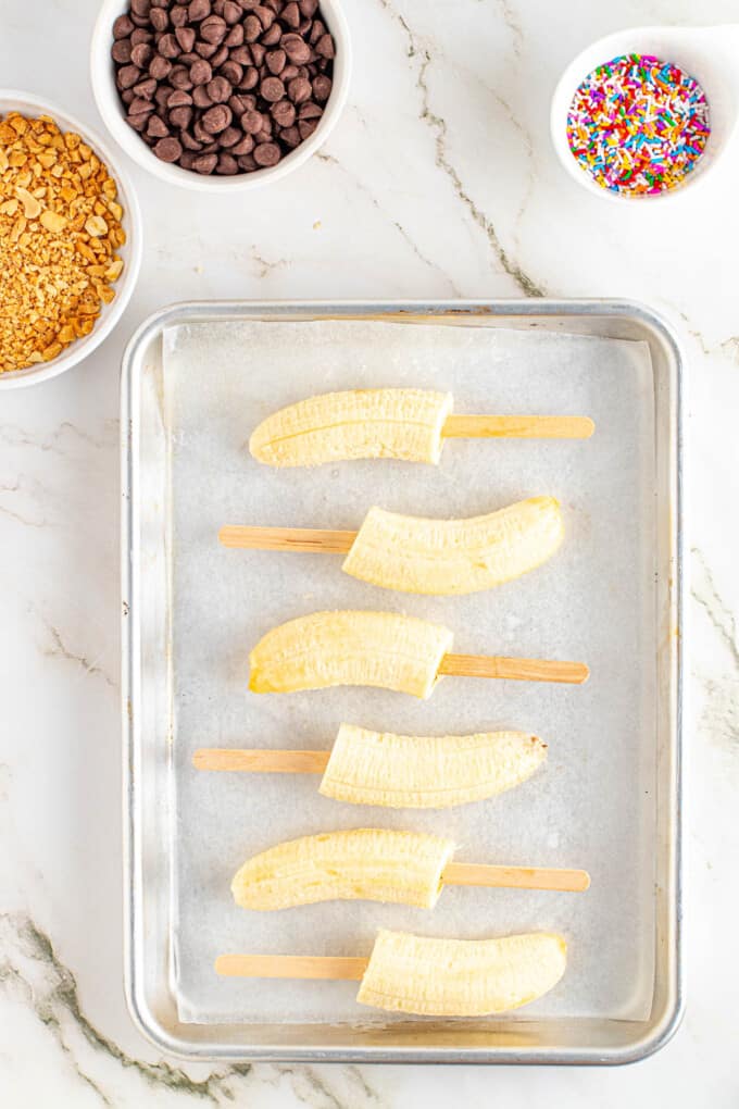 Sticking wooden popsicle sticks into banana halves on white parchment paper.