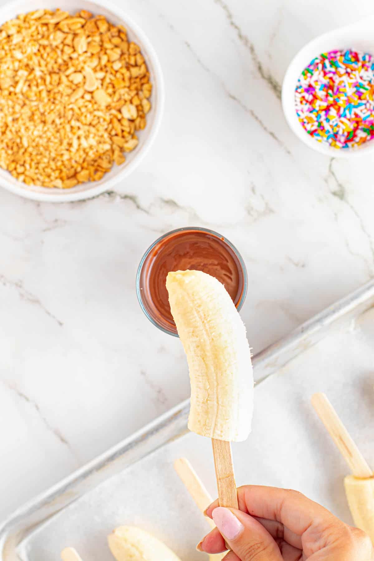 A hand holding a peeled banana half on a wooden popsicle stick.