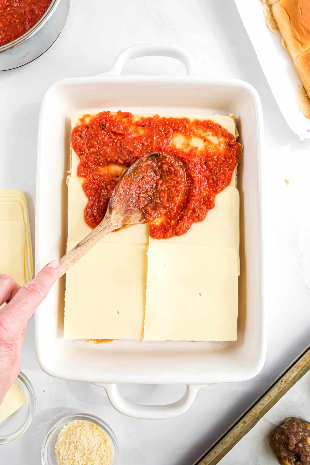 A hand holding a wooden spoon spreading marinara sauce on cheese and buns for meatball sliders.