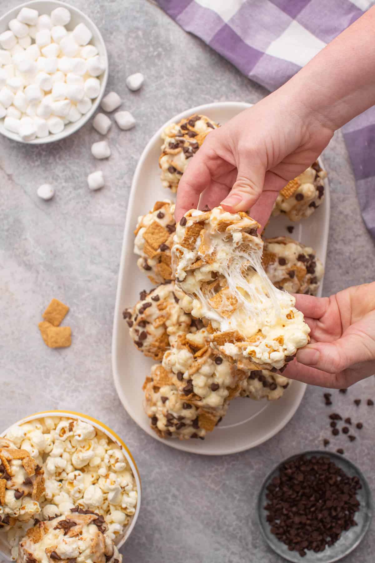 Hands pulling apart a s'mores popcorn ball to showcase the stretchy, gooey marshmallow that holds it together.