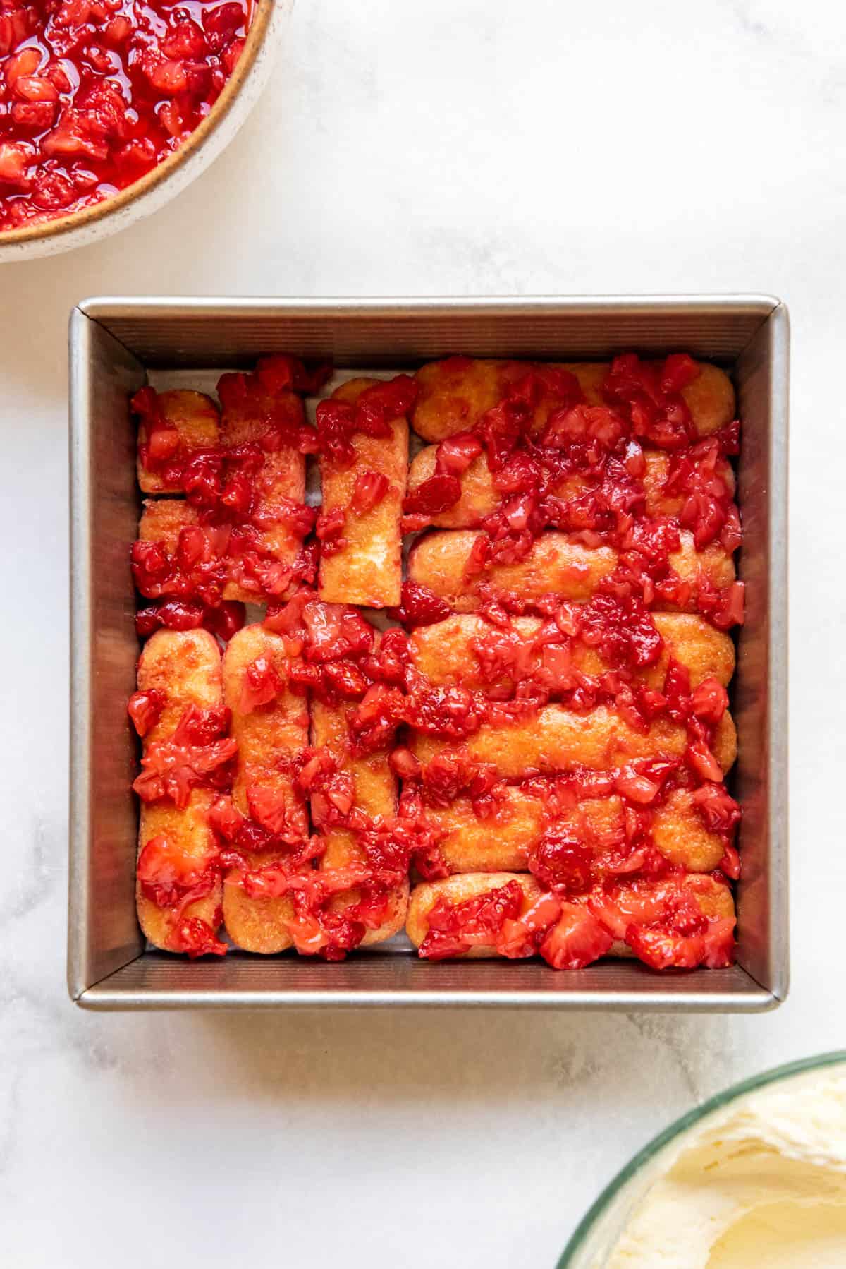 Spreading macerated mashed strawberries over ladyfingers in a pan.