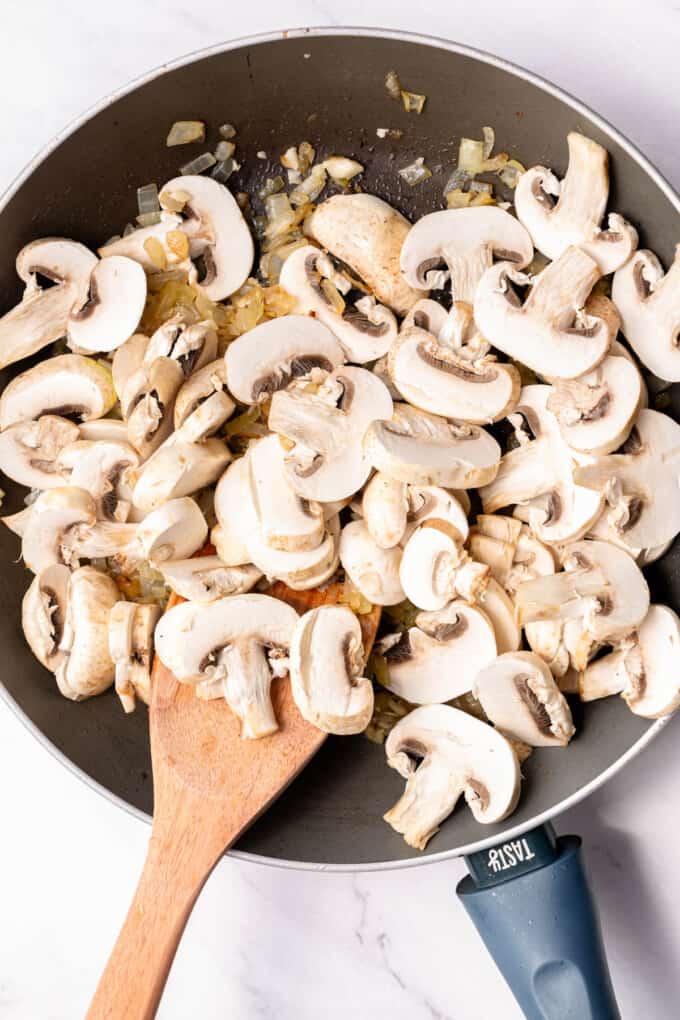 Adding sliced mushrooms to sauteed onions in a large pan. 