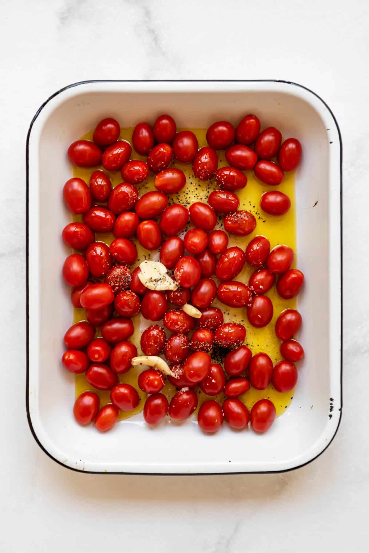 Combining olive oil, grape tomatoes, crushed garlic cloves, and seasoning in a baking dish.
