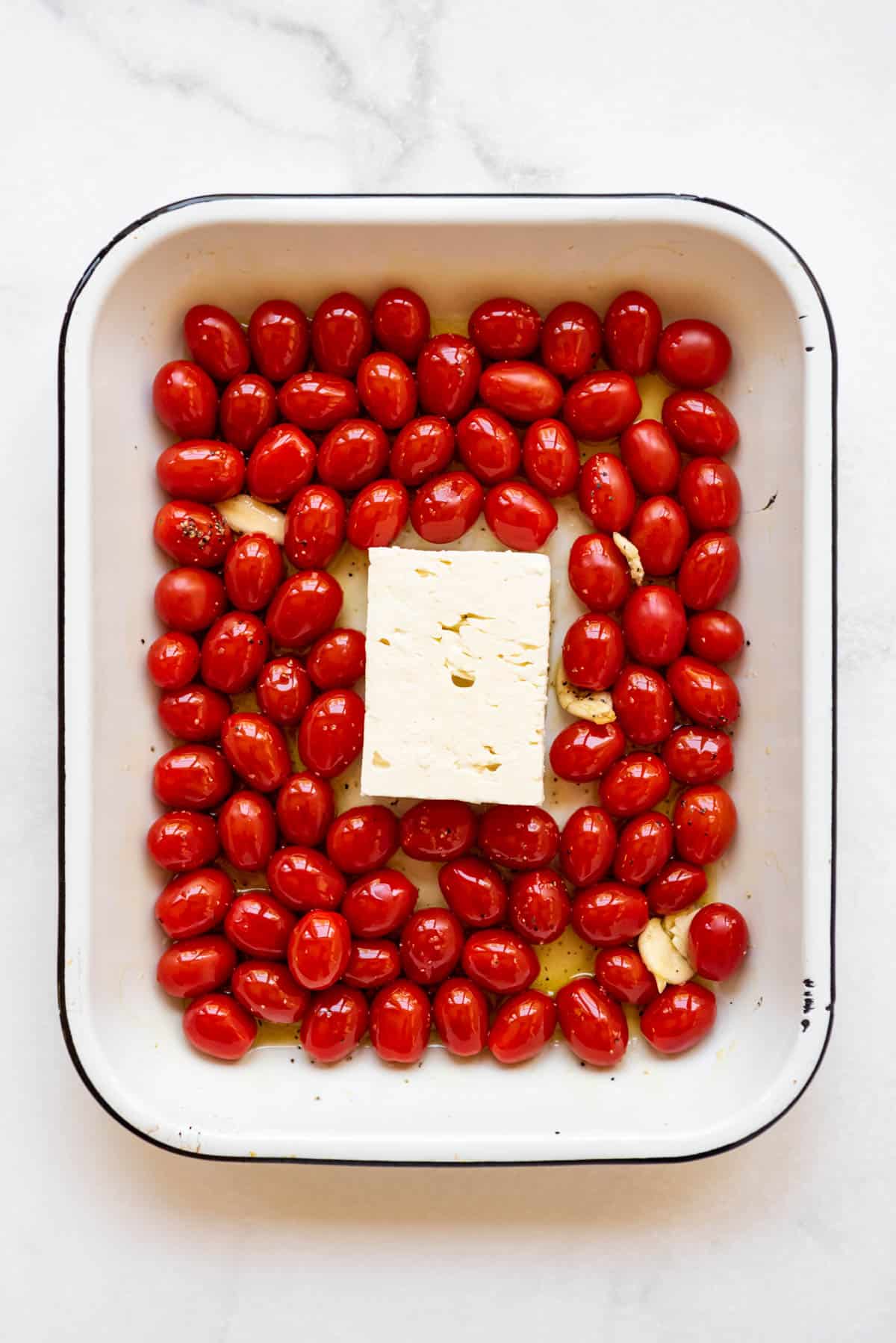 A block of feta cheese nestled in a baking pan filled with grape tomatoes, olive oil, and garlic.