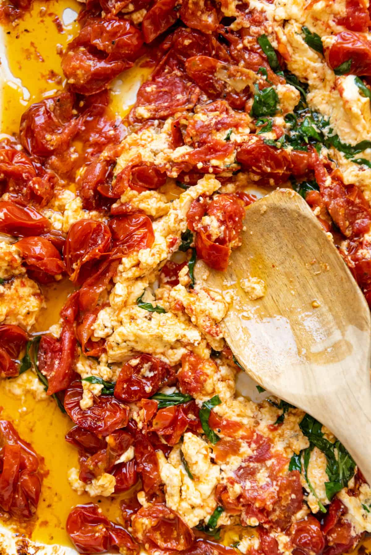 A close image of a wooden spoon mixing together roasted tomatoes and baked feta cheese with olive oil and sliced fresh basil leaves.