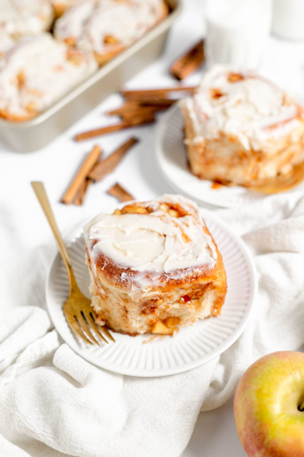 An image of apple cinnamon rolls on white plates next to apples and cinnamon sticks.