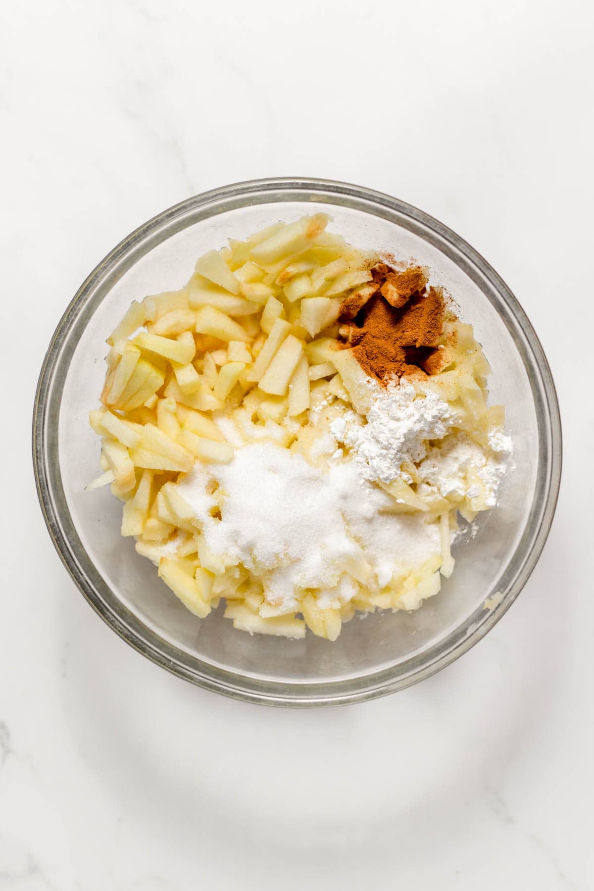 Adding cinnamon, sugar, and flour to chopped, peeled apples in a glass mixing bowl.