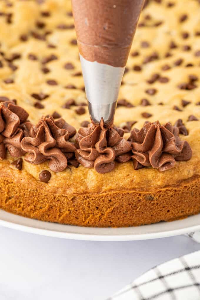 Piping chocolate frosting in a swirl border on top of a chocolate chip cookie cake. 