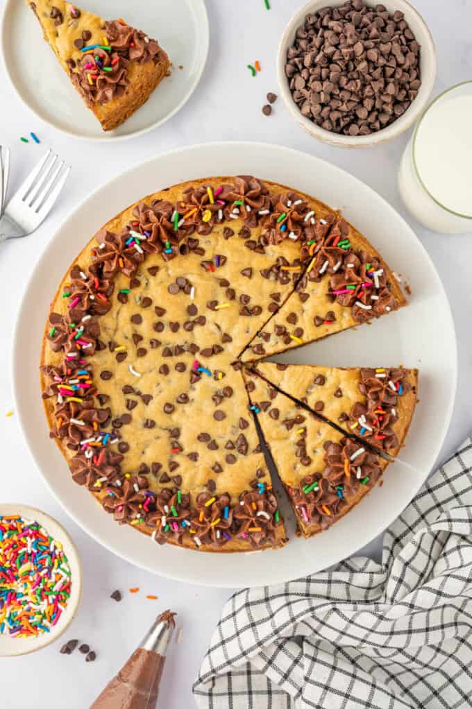 An overhead image of a sliced chocolate chip cookie cake with bowls of mini chocolate chips and sprinkles next to it.