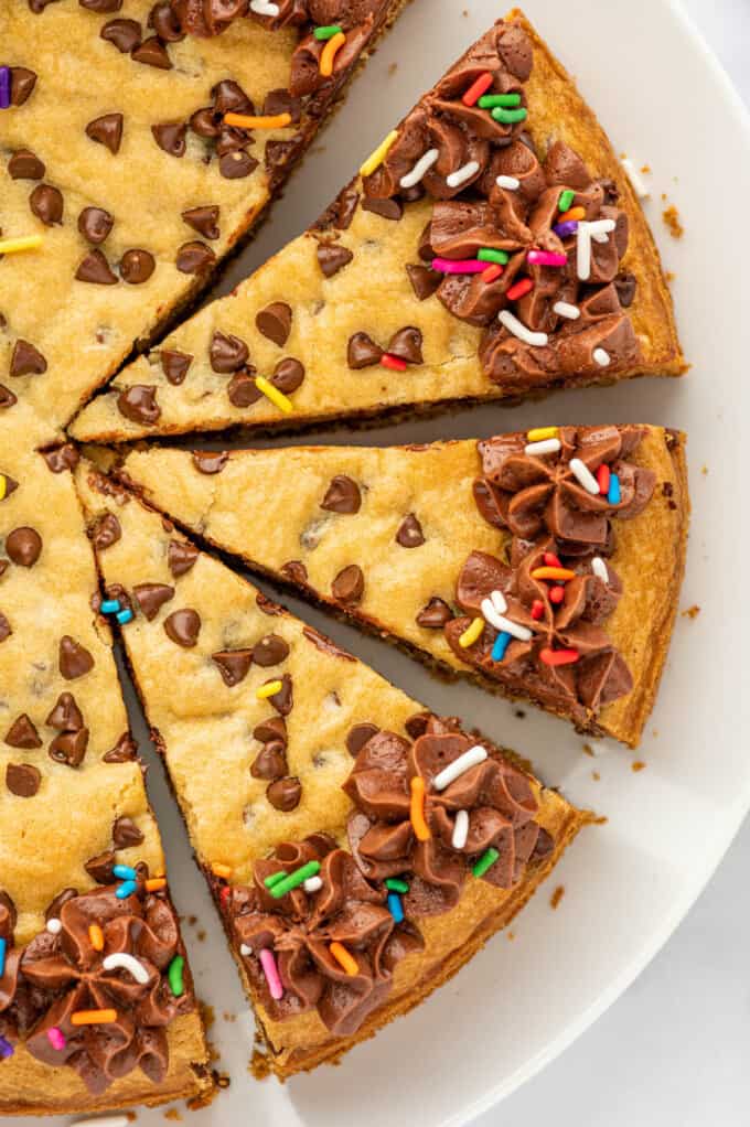 A close image of a sliced chocolate chip cookie cake decorated with frosting and sprinkles.