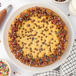 An overhead image of a chocolate chip cookie cake decorated with chocolate frosting and sprinkles.