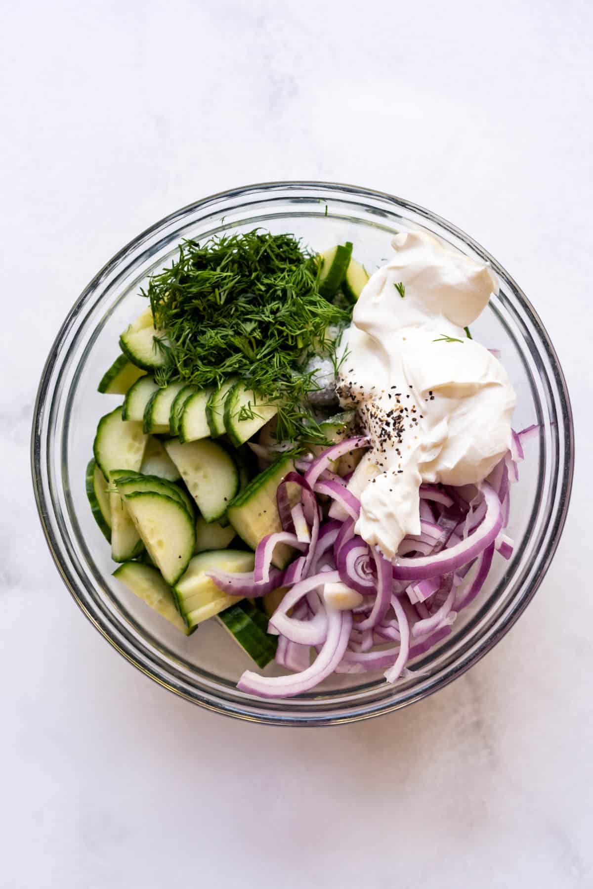 Combining sliced red onions, cucumbers, sour cream, fresh dill, and spices in a bowl to make a cucumber salad.