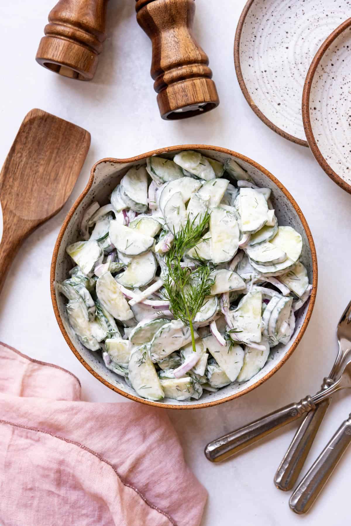 An overhead image of a bowl of creamy German cucumber salad surrounded by a pink cloth napkin, plates, and serving utensils.