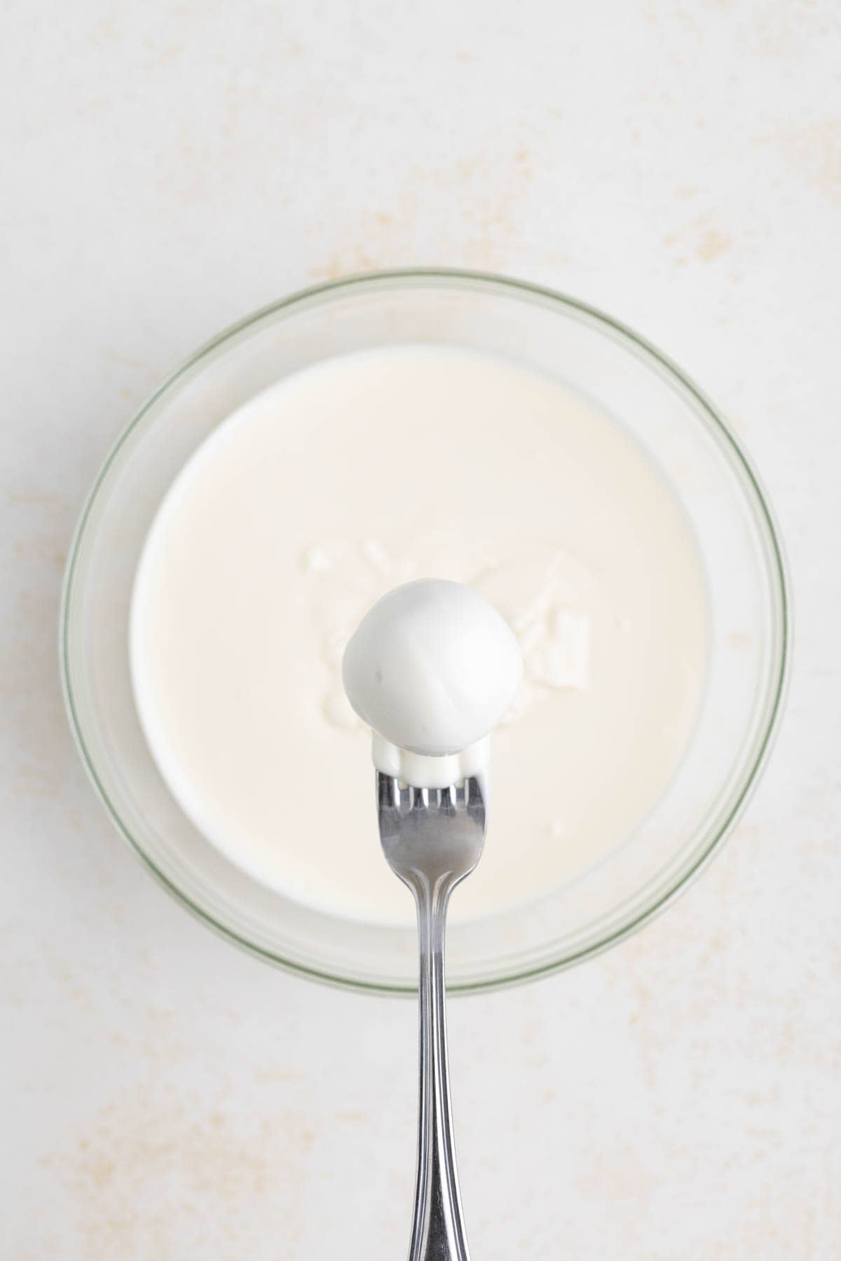 A gingerbread truffle dipped in melted white chocolate being held on a fork over the bowl of white chocolate.